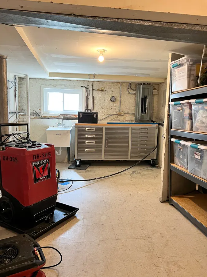A dehumidifier and an air mover drying an unfinished basement with storage shelving, structural drying in Concord, NH